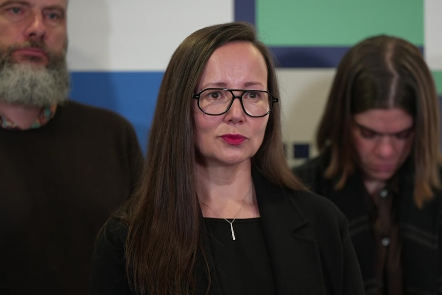 A woman with long dark hair, dark rimmed glasses and a black top stands in front of two people staring off camera.