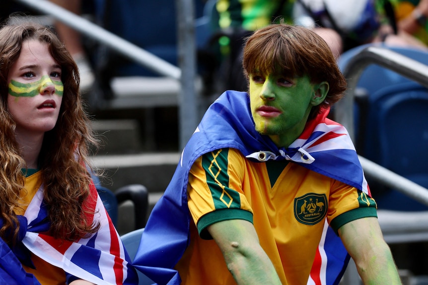 two young people wearing aussie jerseys, australian flags as capes and green and gold face paint