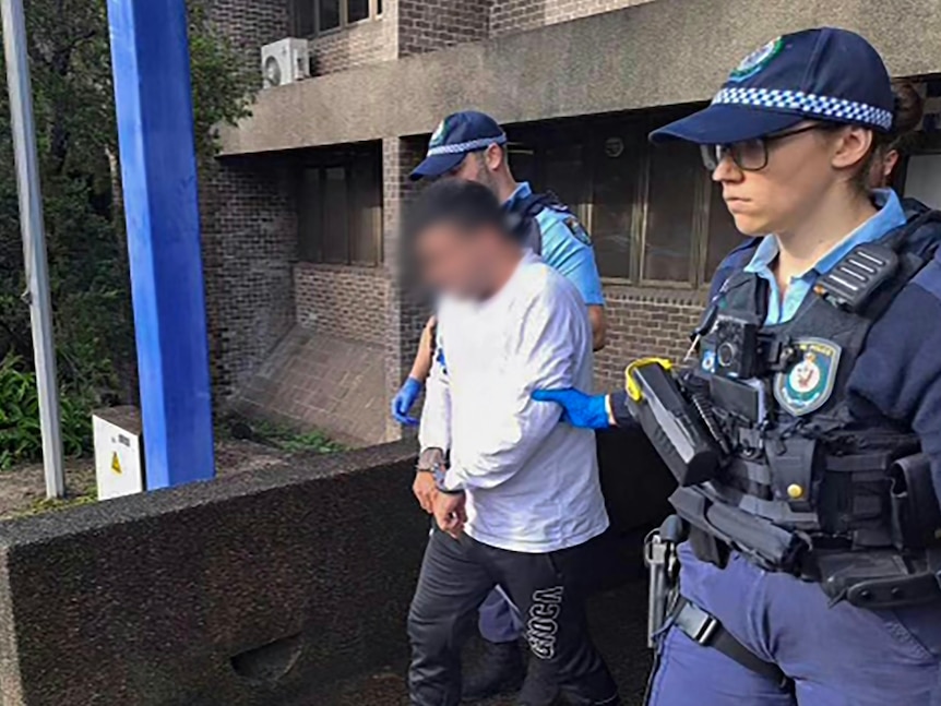 A man in a white long-sleeved t-shirt is escorted by two police officers while in handcuffs outside a police station.