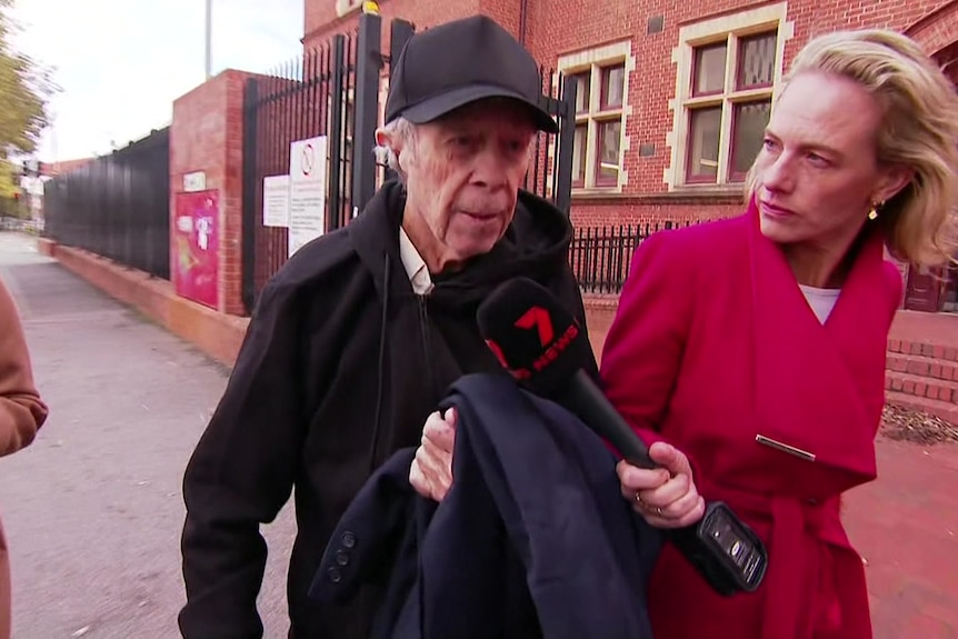 A man in a black jacket and cap is walking along a footpath outside a red brick building with a black fence.