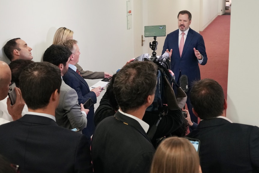 Ed in a suit is questioned by reporters in a hallway