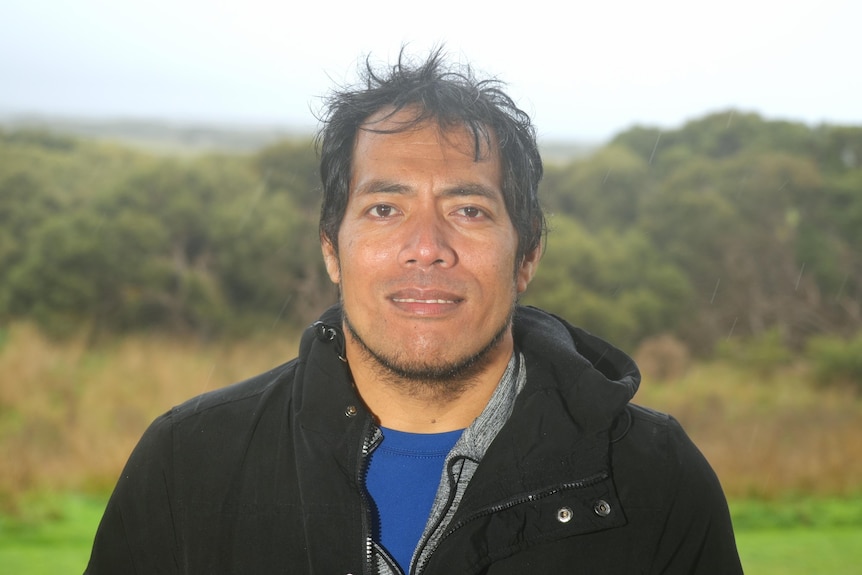 A dark-skinned man wearing a blue t-shirt and a black jacket stands near bushland on a rainy day, smiling at the camera.