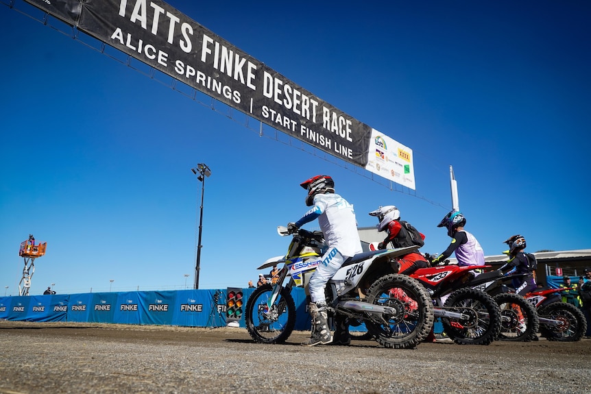 A group of competitors, dressed in riding gear and helmets, revving their motorbikes at the starting line.