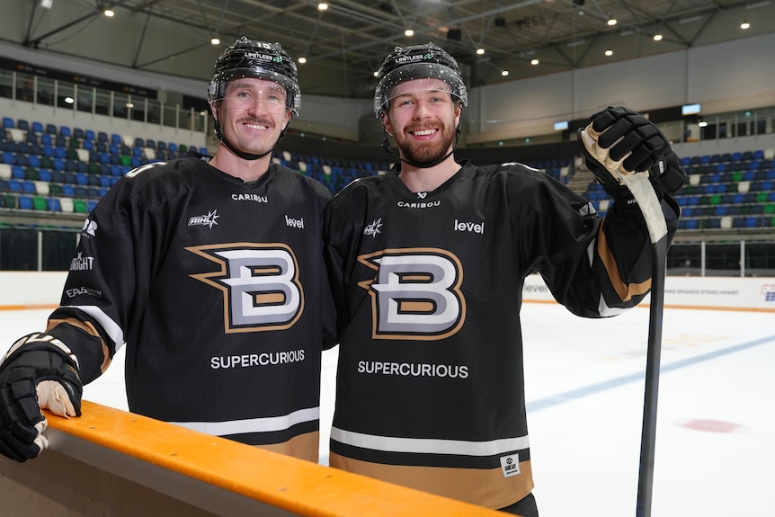 Two people in uniform and helmets holding hockey sticks stand on an ice rink.