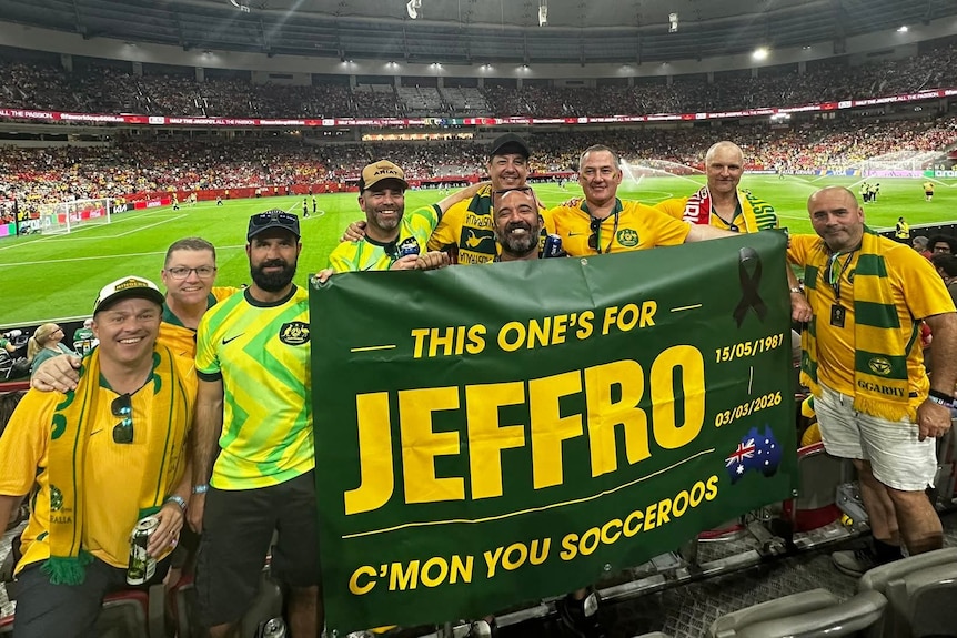 A group of men in jerseys in a sports stadium hold up a banner commemorating a late friend.