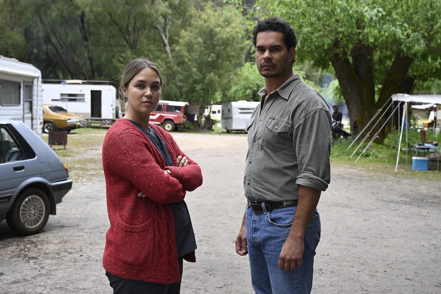 In a caravan park, Tuuli stands next to Mark with her arms crossed as they both look at the camera