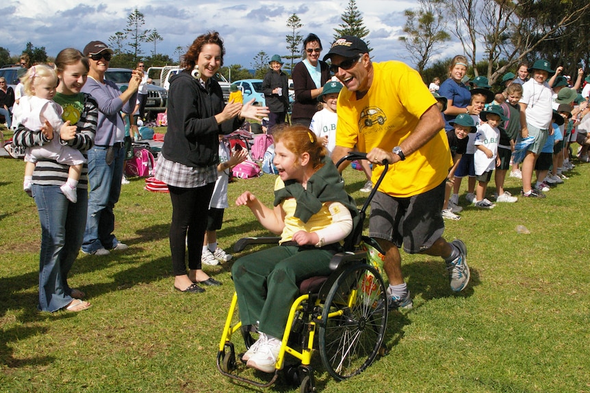 Lillian being pushed along a grass race track in her wheel chair - people lining the edges sheering and clapping her on.