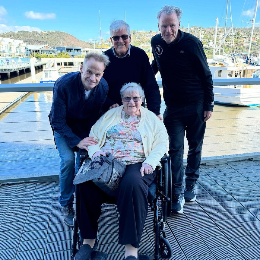 Two adult brothers stand next to their father and lean down towards their mother, who is in a wheelchair, outside sunny day