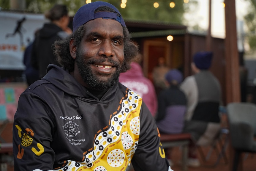 A man in a blue baseball cap and jumper featuring Indigenous designs smiles for the camera.