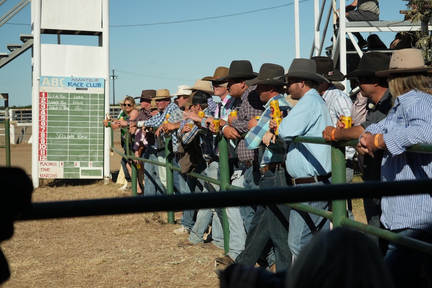 Group of cowboys holding beer cans watch horse racing action 