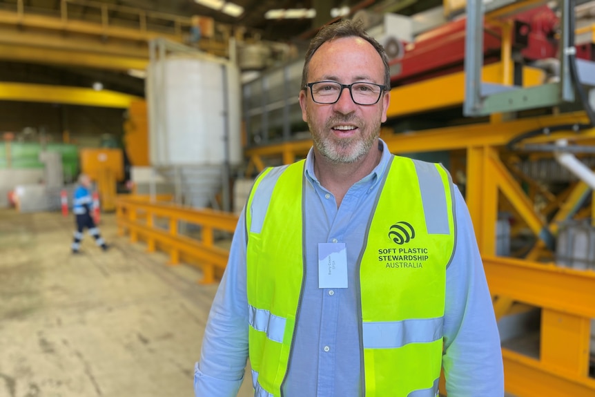 A man in a yellow hi-vis vest stands in a warehouse in front of a large yellow machine.