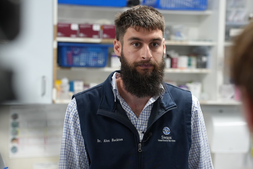 a bearded man in a collared shirt and vest srands in frotn of medicinal cabinet in a vet