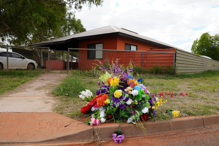 A wide-shot of multi-colour flower memorial resting on the edge of a curb in front of a home, with orange paint.