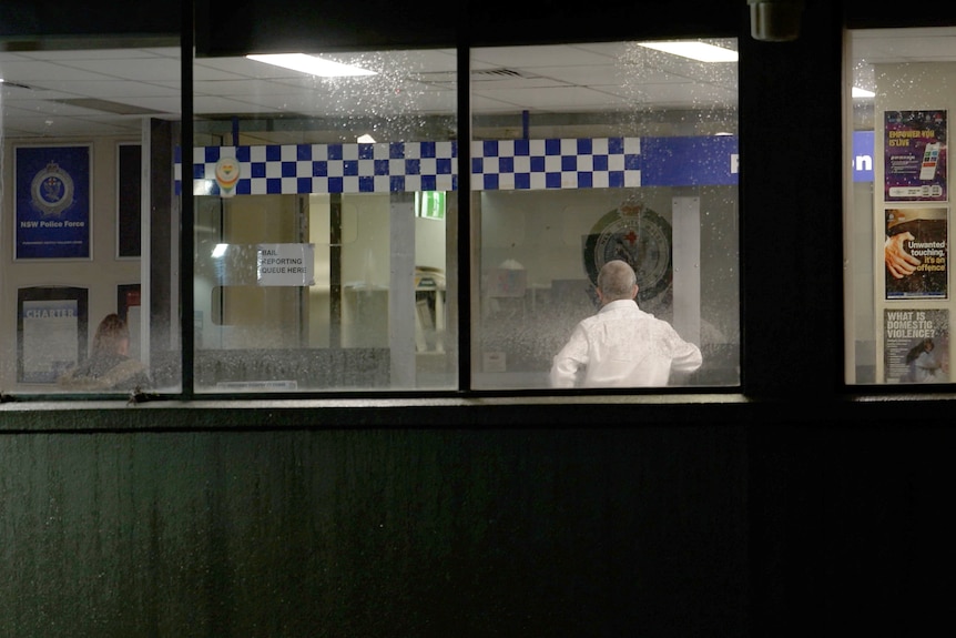 The back of a man wearing a white business shirt standing in a police station is seen through a window.