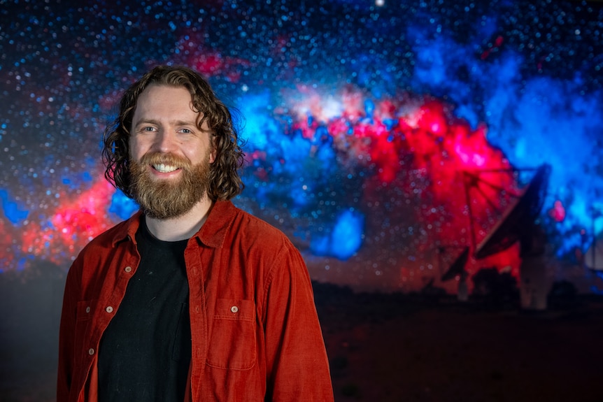 Alec with long hair and beard smiles in front of a silhouette of a radio dish against a generated magnetic field image in sky.