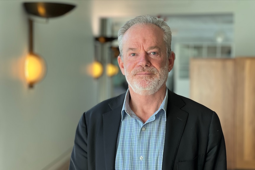 A man with grey hair and a beard, wearing a blue shirt and navy jacket, looking ahead.