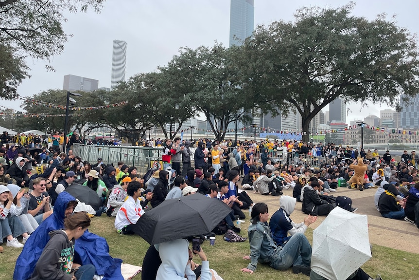 A large crowd of people with some holding umbrellas sit on a grassy area with trees and buildings seen in the distance.