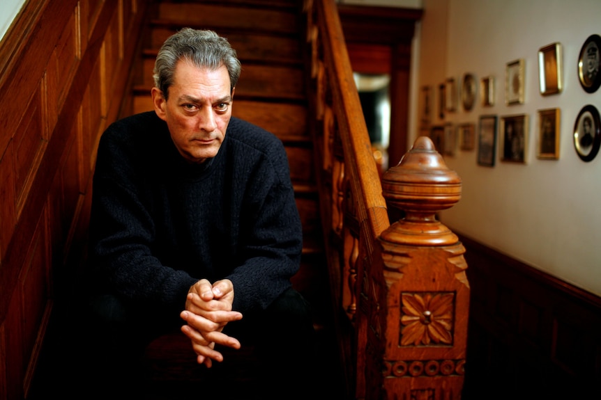 Paul Auster, with greying hair and wearing dark jumper and pants, sitting on a staircase with an ornate timber balustrade.