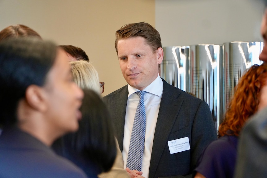 Andrew Hastie stands in a crowd of people wearing a suit and tie with a sticker showing his name.