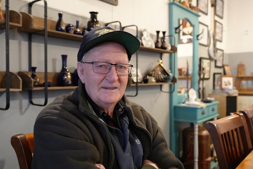 An older man in black sits with his arm folded in a cafe.