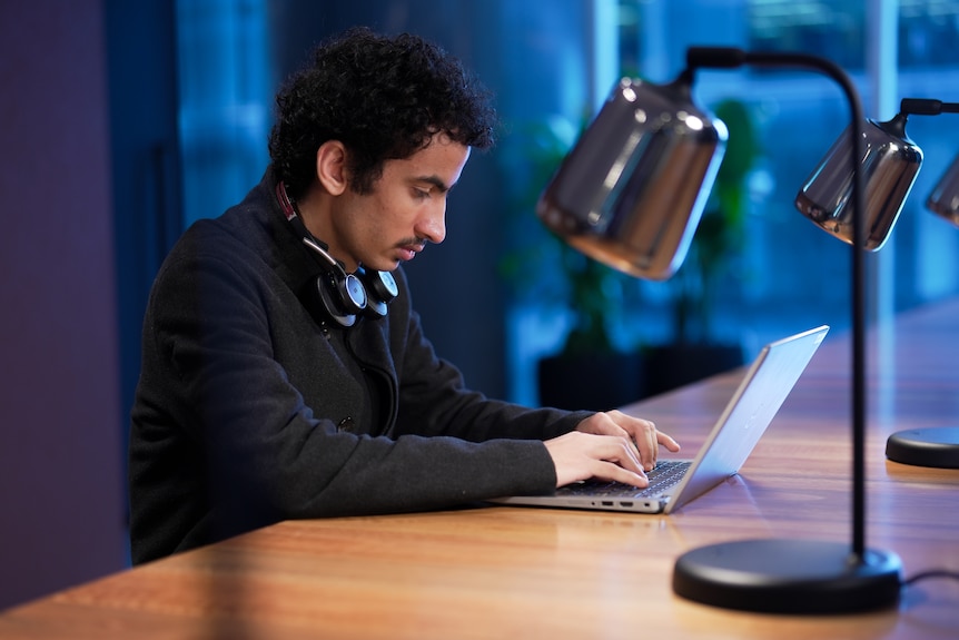 A young man with dark, curly hair sits at a large desk and works on a laptop.