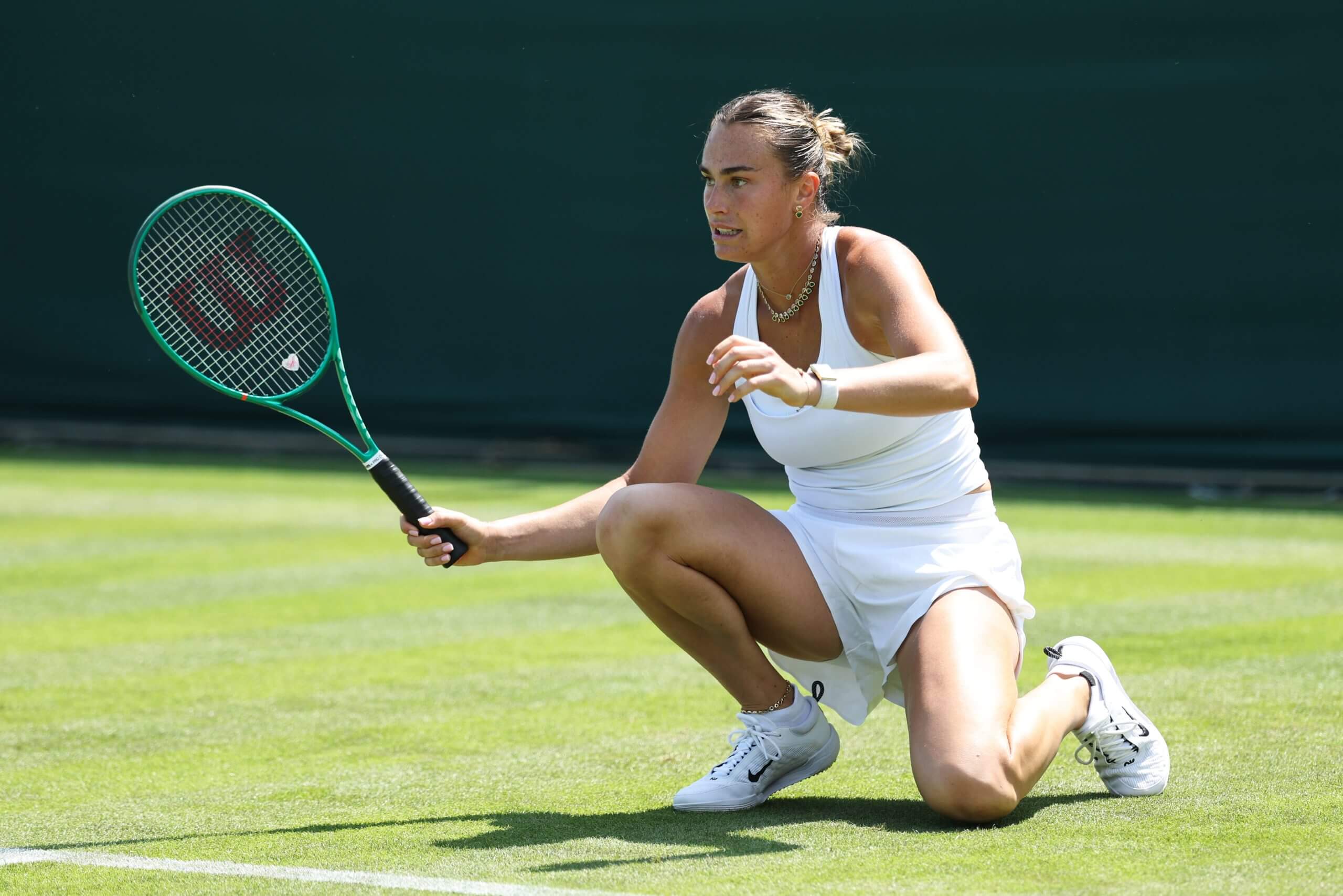 Aryna Sabalenka kneels down on a grass tennis court after hitting a shot.