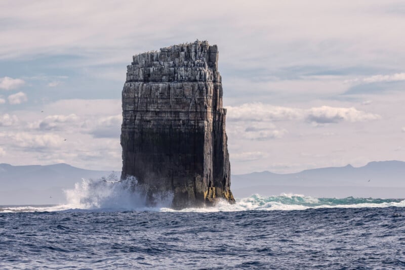 A tall, rocky sea stack rises from the ocean as waves crash against its base, with birds perched on top and a cloudy sky and distant mountains in the background.