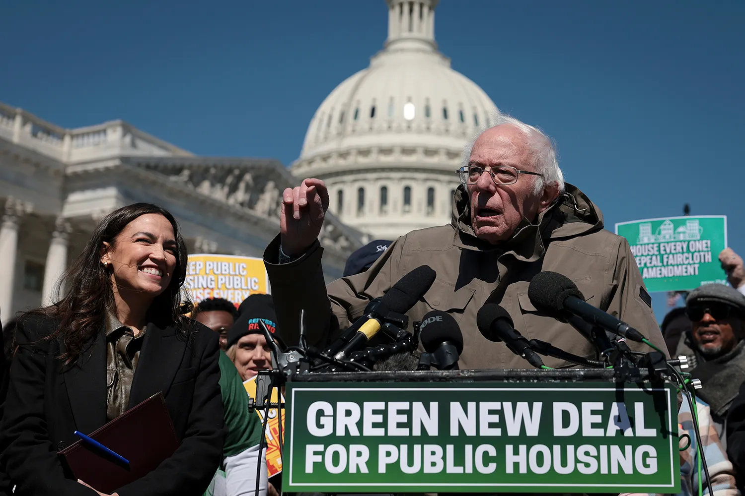 Senator Bernie Sanders speaks forcefully at a podium crowded with microphones, gesturing with his raised right hand. The podium features a prominent green sign that reads "GREEN NEW DEAL FOR PUBLIC HOUSING." To his left, Representative Alexandria Ocasio-Cortez with long dark hair holds a folder and watches him speak. A crowd of people and rally signs are visible in the background, with the large white dome of the U.S. Capitol building rising under a clear blue sky.