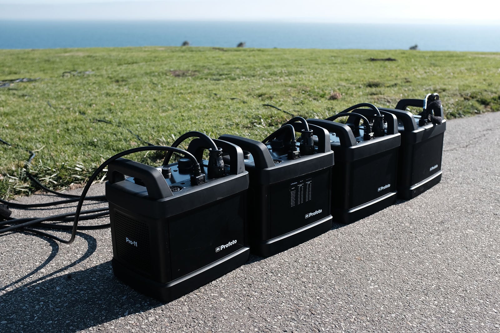 Four black Profoto power packs with cables are lined up on a paved surface outdoors, with green grass and the ocean visible in the background under a clear sky.