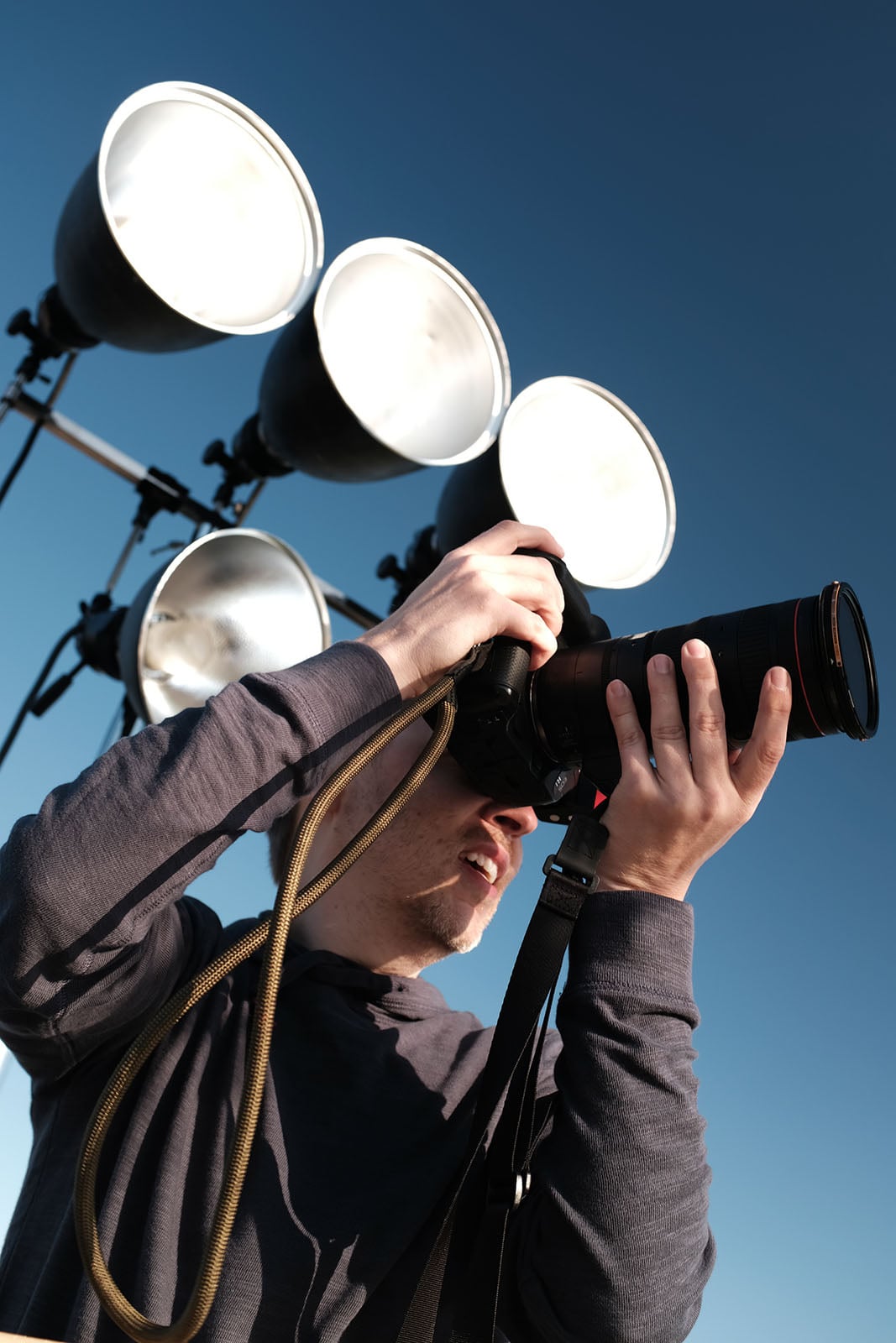 A person holding a camera up to their face, preparing to take a photo outdoors, with four large studio lights visible in the background against a clear blue sky.