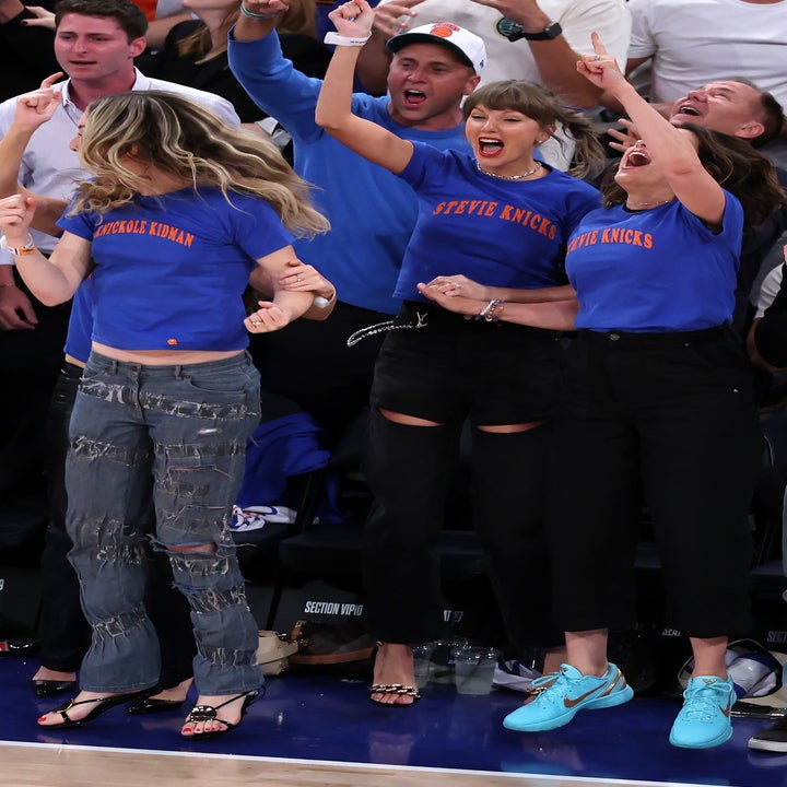 Three women cheer energetically at a sports event, wearing casual outfits with distressed jeans and T-shirts. A lively crowd surrounds them