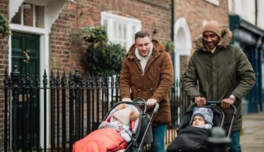 Two male friends are out in Tynemouth, North East UK. They are walking on a sidewalk and pushing their baby sons in strollers. They are wearing warm clothing.