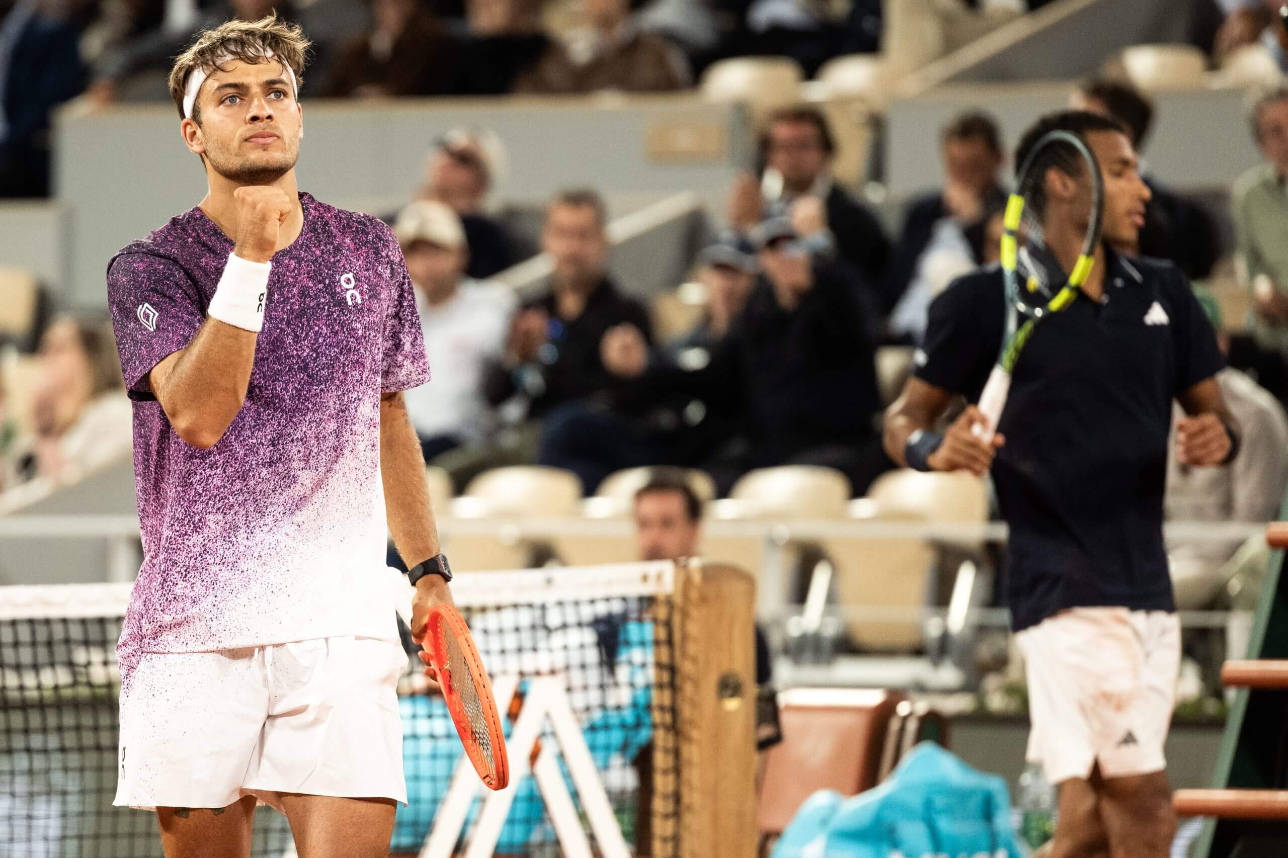 Flavio Cobolli pumps his fist wearing a purple outfit while Félix Auger-Aliassime looks disconsolate to his right.