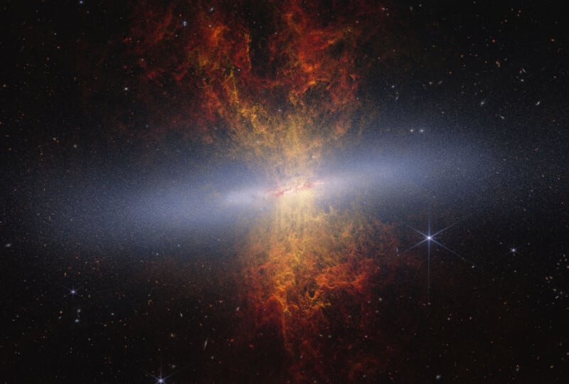 A bright, glowing galaxy with a dense, white core is surrounded by swirling clouds of red and orange gas, set against the dark backdrop of space speckled with stars.
