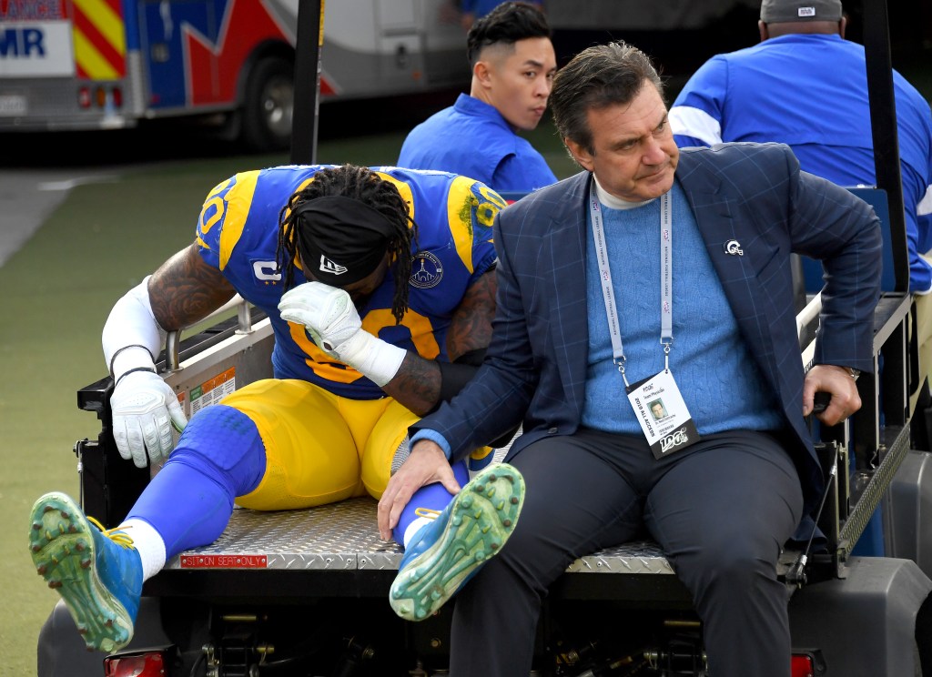 A football player in uniform sits on the back of a cart next to a team physician.