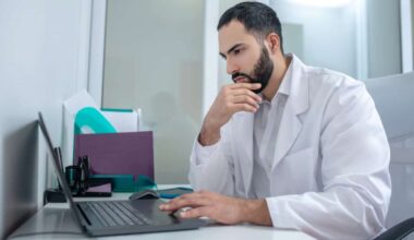 Male doctor in a lab coat working at laptop looking serious.