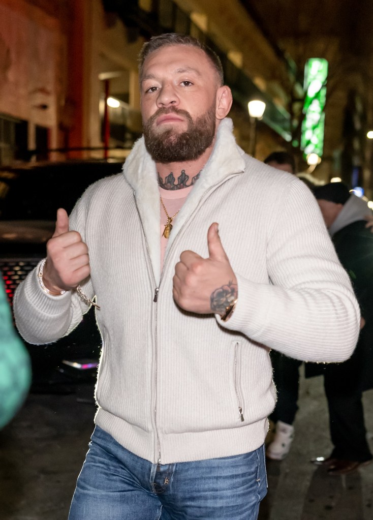Conor McGregor gives two thumbs up while wearing a white ribbed jacket over a pink t-shirt.
