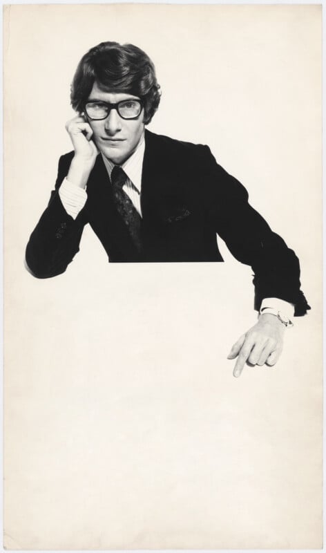 A man in a suit and glasses rests his head on one hand and points downward with the other, against a plain white background.