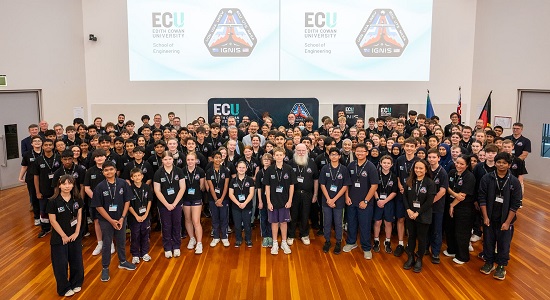 A large group of school students, a few teachers and university lecturers, pose for a photo inside a sports hall