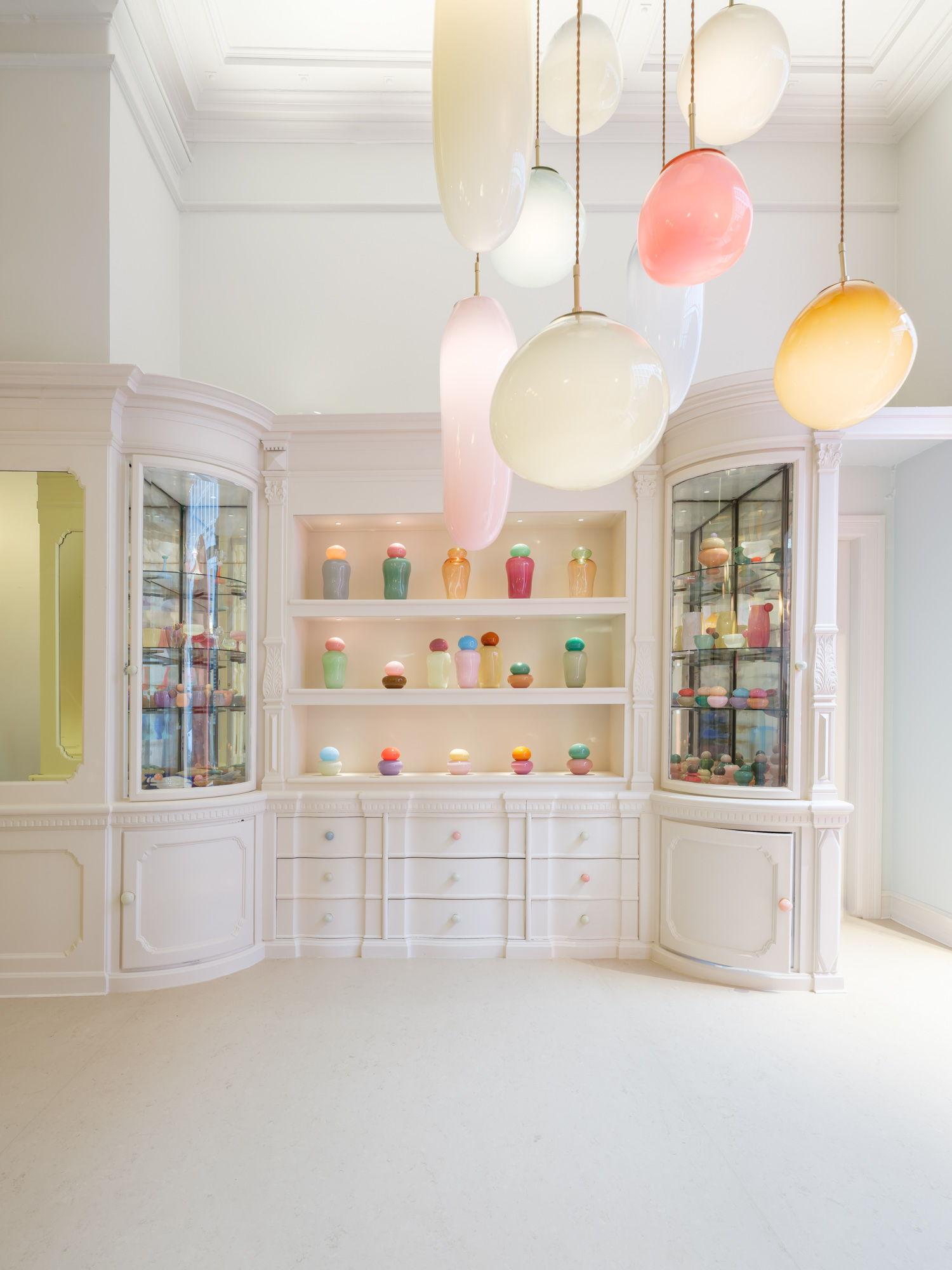 A showroom with white-washed cabinets with colorful, spotty handles, and plenty of mouth-blown glass sculptures on its shelves.