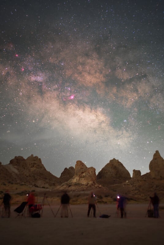 People with tripods and cameras stand in a desert landscape at night, photographing a brightly lit, star-filled sky and the Milky Way above jagged rocky formations.