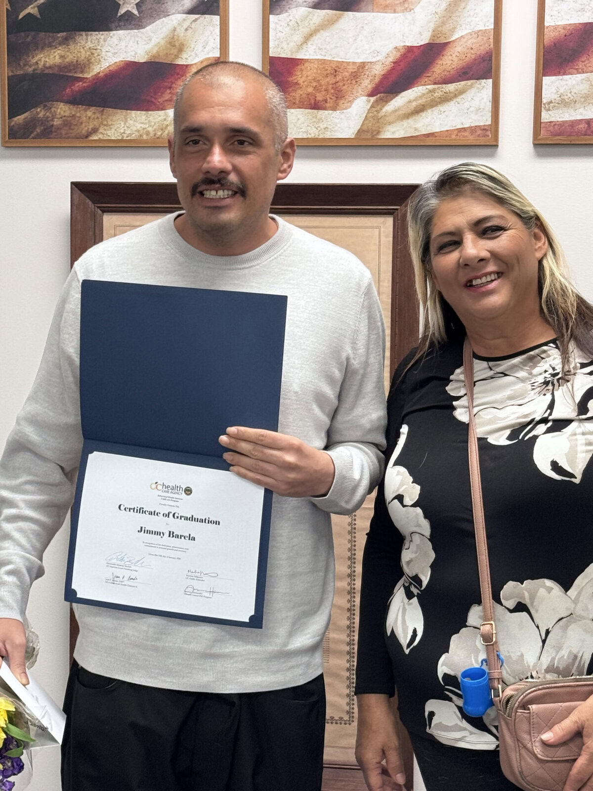 A man holds a graduation certificate. His mom stands next to him.