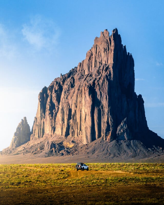 A white SUV parked on a flat, grassy plain with a towering, rugged rock formation rising dramatically in the background under a clear blue sky.