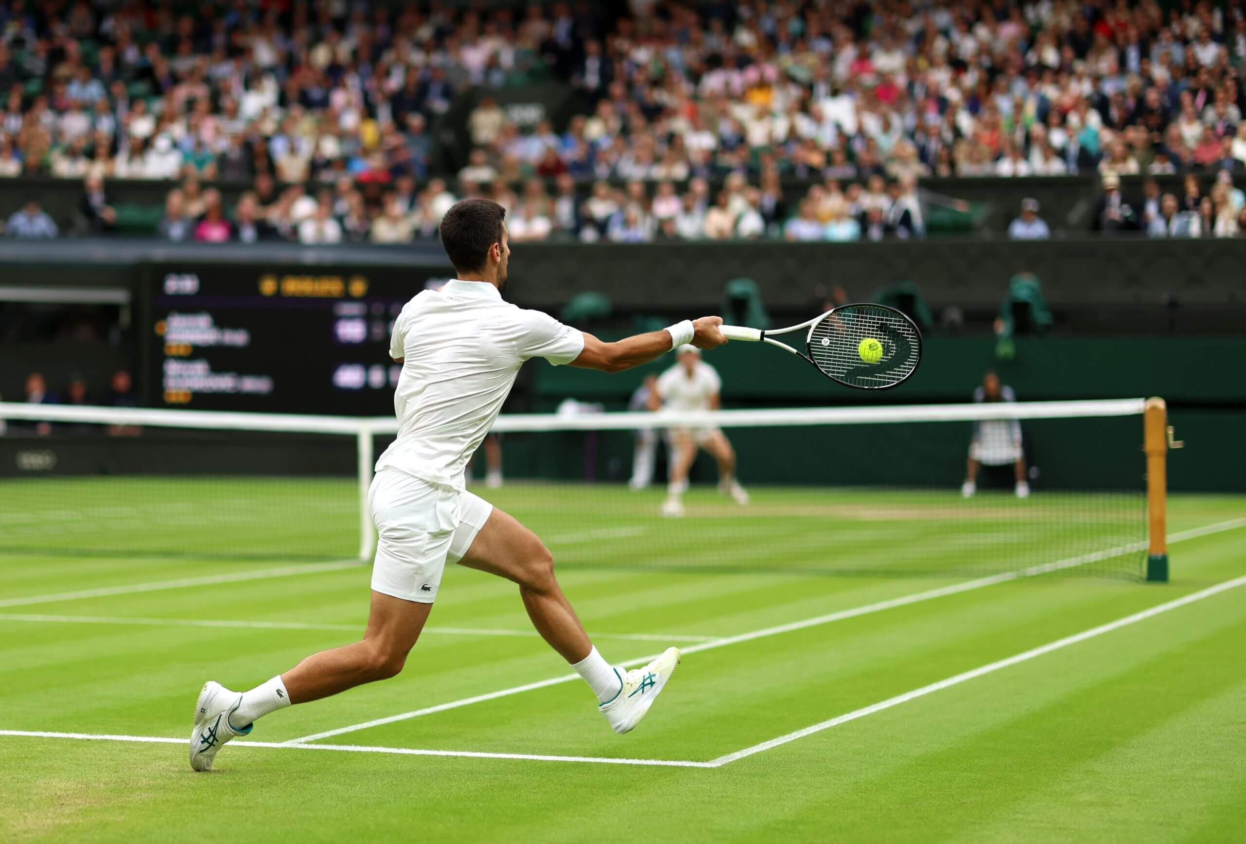 Novak Djokovic hits a forehand on a grass tennis court wearing an all-white outfit, with Jannik Sinner waiting to receive it.