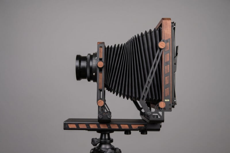 A large format vintage-style camera with a black bellows and wood accents, mounted on a tripod, is shown in profile against a plain gray background.