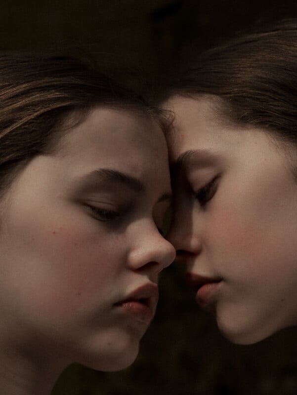 Two young people with fair skin and brown hair gently touch their foreheads and noses together, eyes closed, appearing calm and introspective against a dark, neutral background.