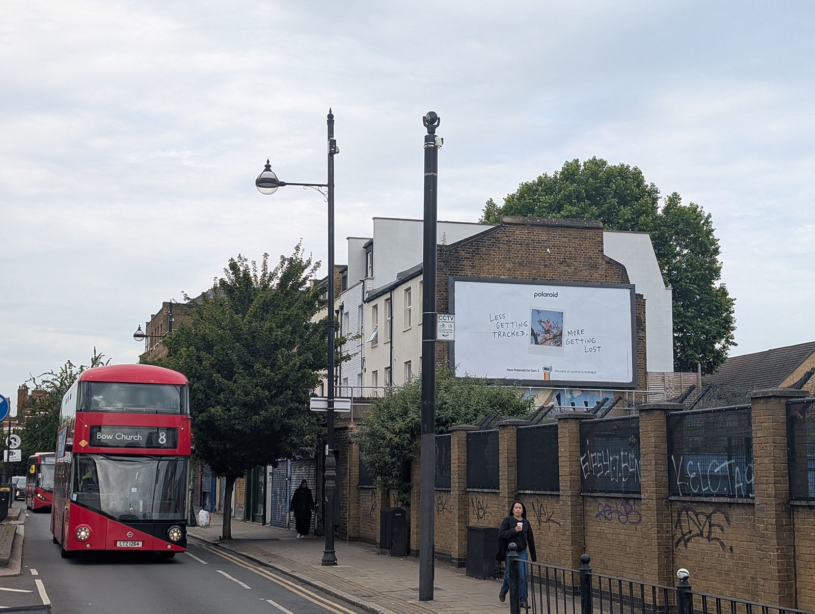 A red double-decker bus labeled "Bow Church" drives down a city street. A billboard on a building reads, "LET’S STRATEGIC TRACKS, NOT GOTTA LIST," with graffiti on the wall below and a woman walking nearby.