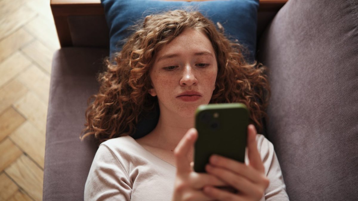 Social media deppression. Sad gen z girl using phone, lying on sofa scrolling on social media, Internet addiction, wasting time. Millennial generation people and gadget use