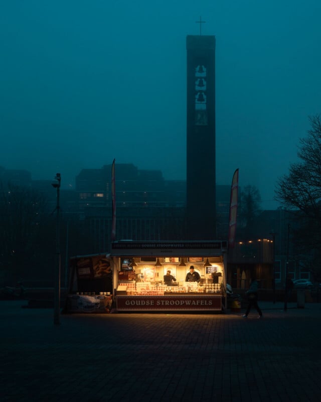 A warmly lit Goudse stroopwafels food stall stands in a foggy square at dusk, with two people inside. A tall, illuminated tower with a cross rises in the background, partially shrouded in mist.