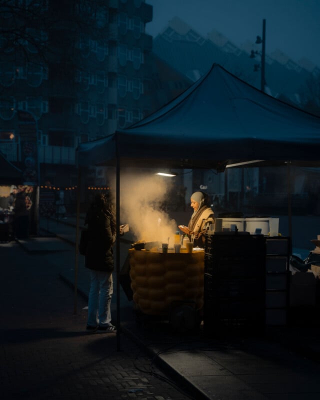 A person stands at an outdoor market stall at dusk, illuminated by warm light and steam rising from a large yellow container, while another person faces them under the canopy, city buildings visible in the background.